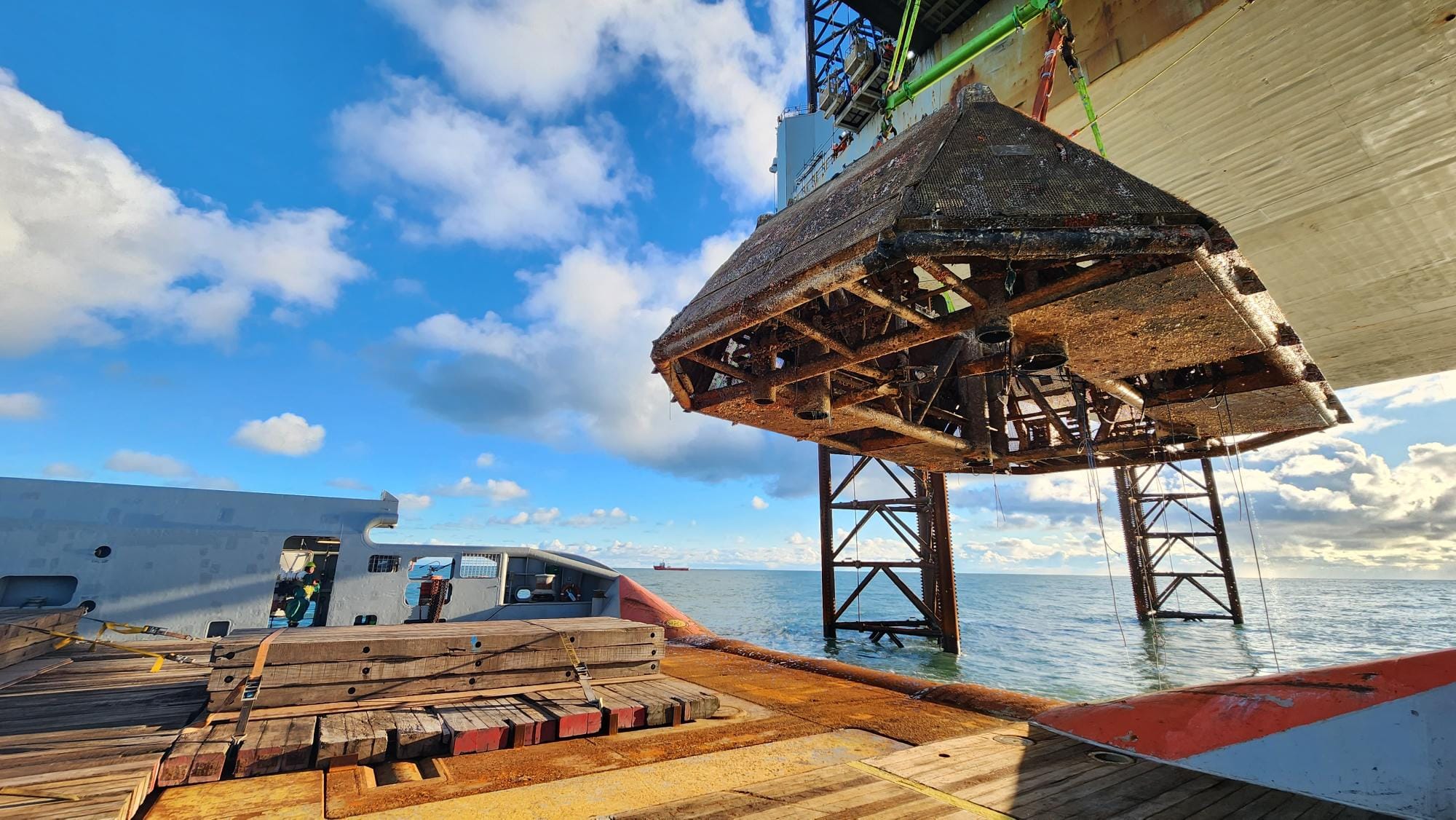 Rusted wellhead protection structure being lifted from the ocean by a crane on an offshore platform.